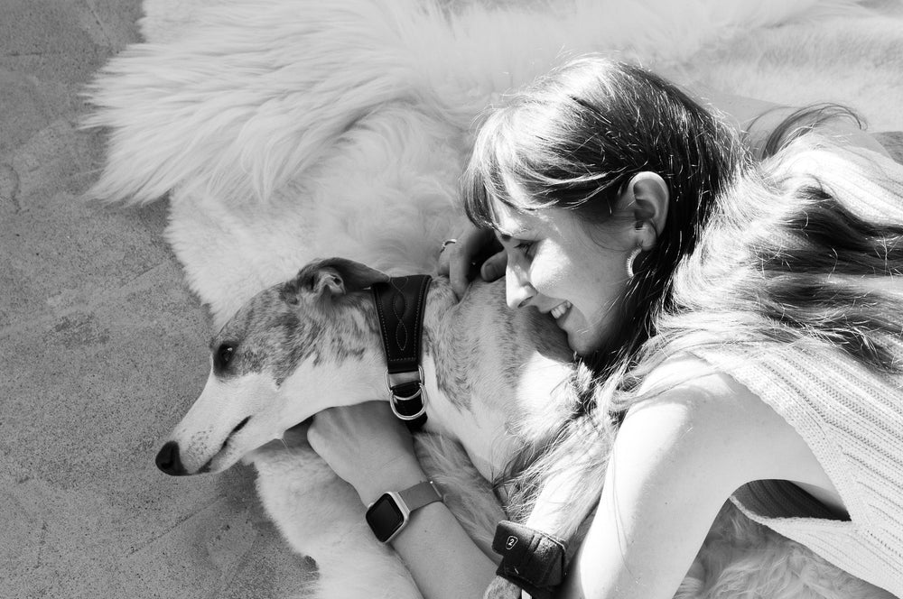 A woman lies next to a relaxed Italian Greyhound on a fluffy white sheepskin rug, both bathed in soft sunlight. She smiles gently while embracing the dog, capturing a quiet moment of connection and comfort.