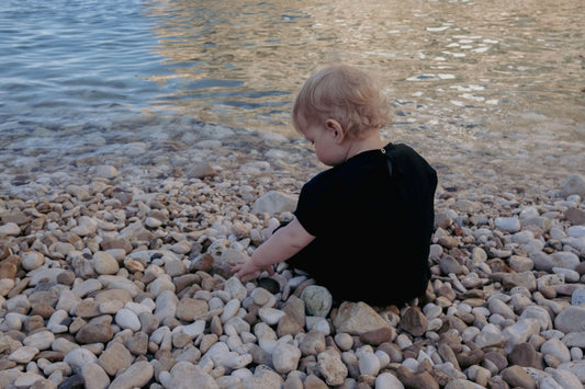 A young child sitting on rocks by a body of water wearing the Ramie Baby/Kid Clothing Set in Charcoal from Mellow Concept Store.