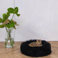 A black cat relaxing on a fluffy Round Natural Sheepskin Pet Bed in front of a potted plant from Mellow Pet Store.